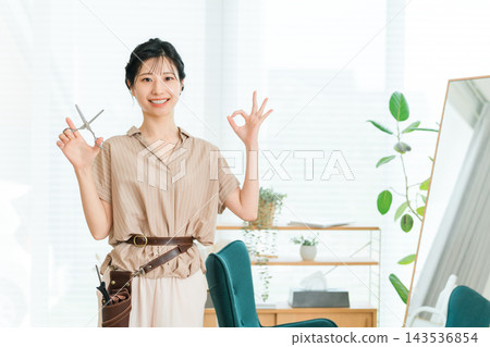 理髮店裡,一位面帶微笑的女理髮師拿著剪刀,比出「OK」手勢。 理髮店裡,一位面帶微笑的女理髮師拿著剪刀,比出「OK」手勢。 143536854