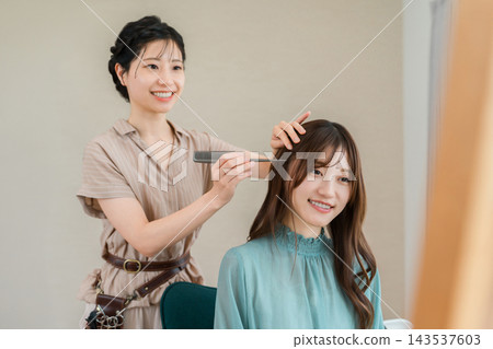 一位微笑的年輕女子正在美容院讓女髮型師為她做頭髮。 一位微笑的年輕女子正在美容院讓女髮型師為她做頭髮。 143537603