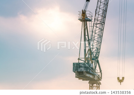 Construction tower crane with a lattice boom, suspended hook and concrete counterweights against a blue and pink sky, heavy lifting equipment for building projects and real estate development. Construction tower crane with a lattice boom, suspended hook and concrete counterweights against a blue and pink sky, heavy lifting equipment for building projects and real estate development. 143538356
