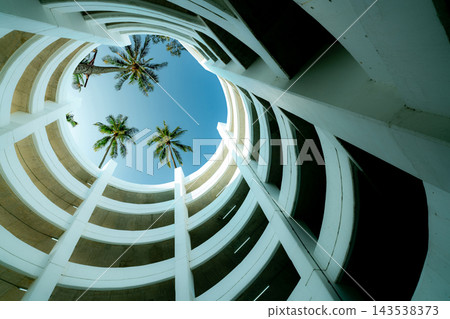Multilevel parking garage with curved concrete floors surrounding a circular opening, white structural columns and coconut palms beneath a blue summer sky, architectural details of a car park. Multilevel parking garage with curved concrete floors surrounding a circular opening, white structural columns and coconut palms beneath a blue summer sky, architectural details of a car park. 143538373