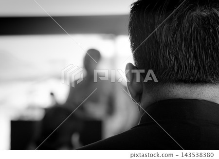 Man wearing a black shirt listening to a monk delivering a sermon at a Buddhist funeral in Thailand, attending a traditional religious ceremony. Man wearing a black shirt listening to a monk delivering a sermon at a Buddhist funeral in Thailand, attending a traditional religious ceremony. 143538380