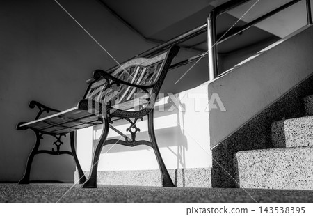 Empty wrought iron bench with wooden slats beside terrazzo stairs in an apartment building, ornate armrests and curved legs beneath a decorative backrest near the metal stair railing. 143538395