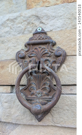 Ornate rusted metal door knocker mounted on textured stone wall, close view of vintage decorative hardware with weathered circular ring, aged finish, and carved floral detail Ornate rusted metal door knocker mounted on textured stone wall, close view of vintage decorative hardware with weathered circular ring, aged finish, and carved floral detail 143540018