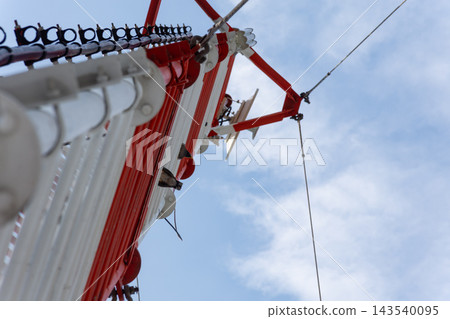 Low angle view of red white communication tower metal antenna structures cables support brackets against blue cloudy sky highlighting wireless transmission telecom infrastructure 143540095