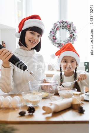 A mother and daughter wearing Santa hats joyfully bake Christmas cookies together in a bright kitchen 143540242