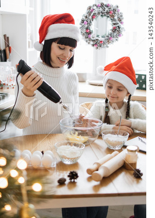 A mother and daughter wearing Santa hats bake together in a festive kitchen preparing holiday treats A mother and daughter wearing Santa hats bake together in a festive kitchen preparing holiday treats 143540243