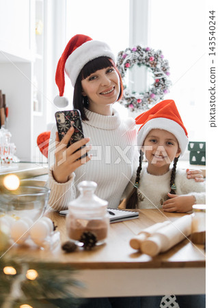 A mother and daughter wearing Santa hats take a selfie while preparing Christmas cookies in a festive kitchen setting 143540244
