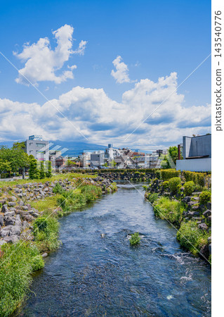 A vertical composition of the clear waters of the Kanda River and the cityscape of Fujinomiya City, Shizuoka Prefecture, against a summery blue sky and cloudy backdrop. A vertical composition of the clear waters of the Kanda River and the cityscape of Fujinomiya City, Shizuoka Prefecture, against a summery blue sky and cloudy backdrop. 143540776