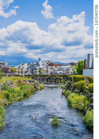 A vertical composition of the clear waters of the Kanda River and the cityscape of Fujinomiya City, Shizuoka Prefecture, against a summery blue sky and cloudy backdrop. A vertical composition of the clear waters of the Kanda River and the cityscape of Fujinomiya City, Shizuoka Prefecture, against a summery blue sky and cloudy backdrop. 143540786