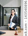 Asian female lawyer in a suit holding a contract in her office. Confident attorney standing by a desk Asian female lawyer in a suit holding a contract in her office. Confident attorney standing by a desk 143540896