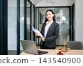 Asian female lawyer in a suit holding a contract in her office. Confident attorney standing by a desk Asian female lawyer in a suit holding a contract in her office. Confident attorney standing by a desk 143540900