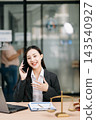 Asian female lawyer talking on a smartphone at her modern office desk. Confident attorney giving legal advice near a laptop, gavel Asian female lawyer talking on a smartphone at her modern office desk. Confident attorney giving legal advice near a laptop, gavel 143540927