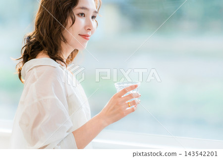 A woman smiles, holding a glass, by the window in her living room. A relaxing time bathed in natural light. A woman smiles, holding a glass, by the window in her living room. A relaxing time bathed in natural light. 143542201