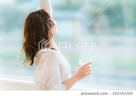 一位女士在客廳窗邊放鬆，手裡拿著玻璃杯，伸展著雙臂。 143542203