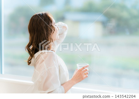 一位女士在客廳的窗邊放鬆地坐著，手裡拿著一個玻璃杯，眺望著窗外。 143542204