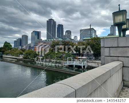 A dark river reflecting low-hanging clouds, brick buildings and green trees beyond the pier, with a backdrop of skyscrapers. A dark river reflecting low-hanging clouds, brick buildings and green trees beyond the pier, with a backdrop of skyscrapers. 143543257