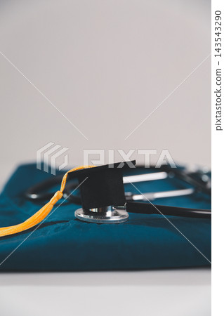Graduation cap resting on a stethoscope over blue medical scrubs, symbolizing medical graduation, healthcare education, academic achievement and a professional healthcare career. Graduation cap resting on a stethoscope over blue medical scrubs, symbolizing medical graduation, healthcare education, academic achievement and a professional healthcare career. 143543290