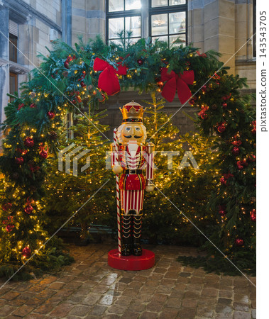 Festive Nutcracker Figure Among Glowing Christmas Trees. A large red and white nutcracker figure with a golden crown stands among glowing Christmas trees decorated with red bows, baubles and fairy lig 143543705