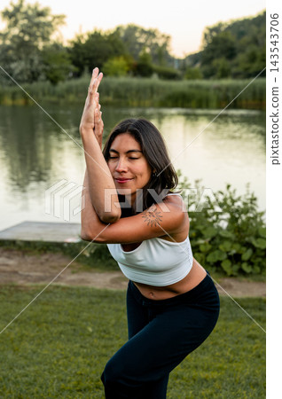 Young Woman in Standing Eagle Twist on a Dock by the Lake. A young Hispanic woman practises a standing eagle twist on a wooden dock over the calm lake at sunset, a wellness routine for physical and me Young Woman in Standing Eagle Twist on a Dock by the Lake. A young Hispanic woman practises a standing eagle twist on a wooden dock over the calm lake at sunset, a wellness routine for physical and me 143543706