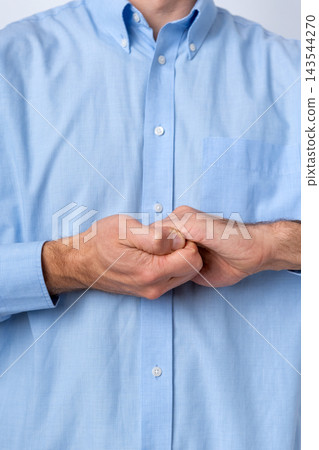 Man's hands clasped tightly together in front of his torso, wearing a light blue dress shirt, conveying concepts of focus, patience, anticipation, or contemplation in a professional setting 143544270