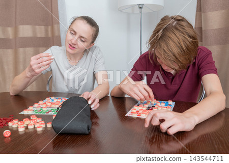 Adult woman and young boy focus on bingo cards at a wooden table in a cozy home, enjoying family game night with tokens, numbers and friendly competition and bonding Adult woman and young boy focus on bingo cards at a wooden table in a cozy home, enjoying family game night with tokens, numbers and friendly competition and bonding 143544711