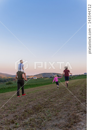 Family walking together up a grassy hill during sunset, father carrying a young boy on his shoulders while two other children run ahead, symbolizing togetherness and outdoor recreation Family walking together up a grassy hill during sunset, father carrying a young boy on his shoulders while two other children run ahead, symbolizing togetherness and outdoor recreation 143544712