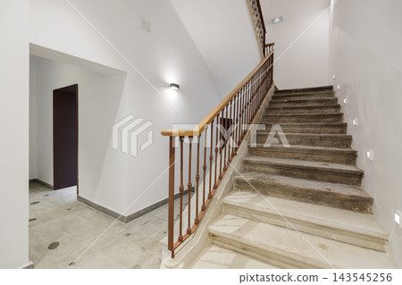 A modern staircase in a well-lit hallway. The stairs are made of stone with a wooden railing. The walls are painted white, creating a clean and minimalist look. 143545256