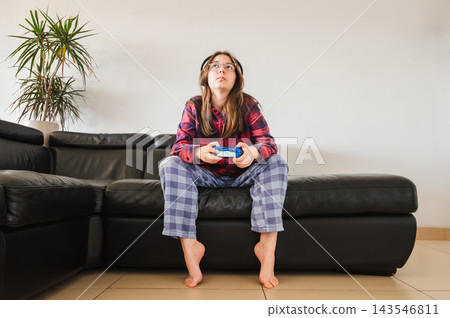 Full Length View of a Teen Girl Gaming at Home. A full length view of a teenage girl sitting on the edge of the couch, looking up at the screen as she plays a video game at home. 143546811