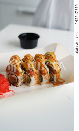 Sushi rolls with colorful sauces, fried toppings, and pickled ginger in an open takeaway box on a white table Sushi rolls with colorful sauces, fried toppings, and pickled ginger in an open takeaway box on a white table 143547856