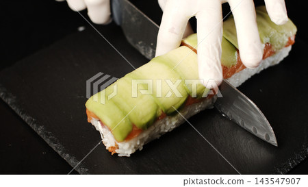 Chef's gloved hands slicing a sushi roll featuring fresh avocado, salmon, and sticky rice on a dark slate board 143547907