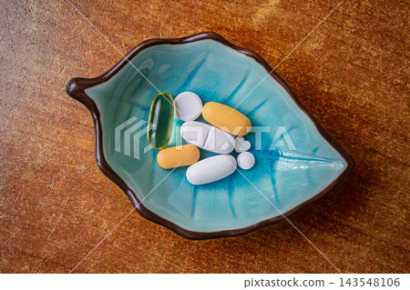 Assorted pills in a turquoise leaf shaped ceramic dish on a wooden table. Assorted pills in a turquoise leaf shaped ceramic dish on a wooden table. 143548106