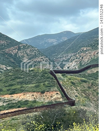 The landscape of the border wall between Mexico and the United States, winding through the rugged, green mountainous region of Baja California. 143552246