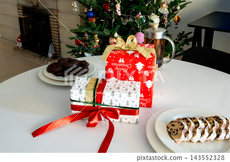 Red and Snowflake Gifts with Stollen by Christmas Tree. A red Merry Christmas gift and a black snowflake present beside stollen and chocolate cookies on a white table before a glowing Christmas tree Red and Snowflake Gifts with Stollen by Christmas Tree. A red Merry Christmas gift and a black snowflake present beside stollen and chocolate cookies on a white table before a glowing Christmas tree 143552328
