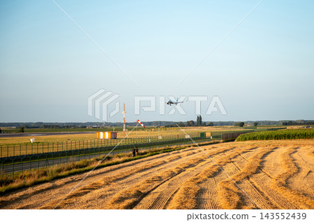 Distant Airliner Taking Off Over a Field at Sunset. A passenger jet climbs into the sky far beyond a harvested field at Brussels Airport in Belgium, the small silhouette of the aircraft in warm golden Distant Airliner Taking Off Over a Field at Sunset. A passenger jet climbs into the sky far beyond a harvested field at Brussels Airport in Belgium, the small silhouette of the aircraft in warm golden 143552439