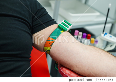 Donor Arm with Colorful Tourniquet on Donation Chair. Close-up of a blood donor's arm with a colorful tourniquet strap resting on a red donation chair beside sample tubes, illustrating preparation Donor Arm with Colorful Tourniquet on Donation Chair. Close-up of a blood donor's arm with a colorful tourniquet strap resting on a red donation chair beside sample tubes, illustrating preparation 143552463