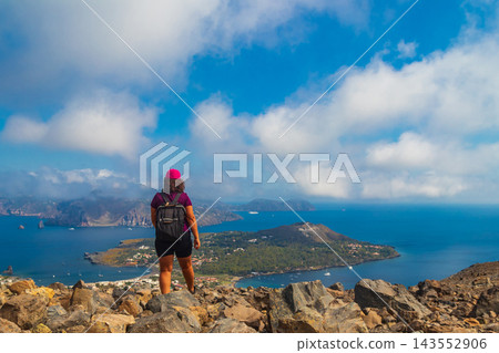 Woman hiker with a backpack standing on top of a volcano mountain, enjoying the scenic view of the blue Tyrrhenian sea, Vulcanello peninsula and Lipari island in the Aeolian archipelago, Sicily, Italy Woman hiker with a backpack standing on top of a volcano mountain, enjoying the scenic view of the blue Tyrrhenian sea, Vulcanello peninsula and Lipari island in the Aeolian archipelago, Sicily, Italy 143552906