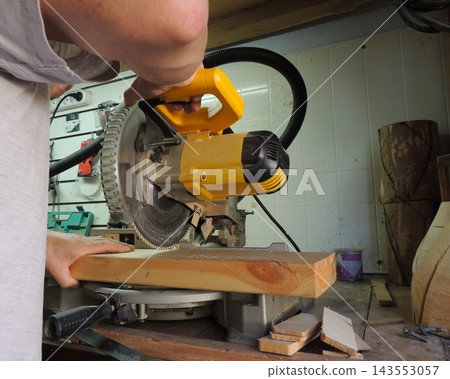 A man lowers the handle of a running yellow miter saw to cut through a thick wooden beam. His other hand firmly holds the workpiece in place on the base. A close-up of the woodworking process. A man lowers the handle of a running yellow miter saw to cut through a thick wooden beam. His other hand firmly holds the workpiece in place on the base. A close-up of the woodworking process. 143553057