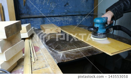A wood processing scene in a workshop. The hand of a craftsman wearing a leather jacket guides a blue sander, smoothing the surface of a board. Finished wooden blocks lie on the workbench nearby. A wood processing scene in a workshop. The hand of a craftsman wearing a leather jacket guides a blue sander, smoothing the surface of a board. Finished wooden blocks lie on the workbench nearby. 143553058