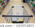 First-person view of a metallic shopping cart moving through a supermarket aisle between shelves stocked with products 143553855