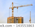 Yellow tower crane stands above a multi-story residential building under construction against a clear blue sky, lifting heavy machinery and materials for urban estate development 143553863