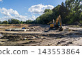 Leveled construction site featuring piles of excavated soil and excavators. Construction machinery clears a plot of land surrounded by a green forest under a cloudy sky 143553864
