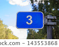 Blue rectangular metal road sign displaying white number three stands mounted on metal pole outdoors. Navigation pointer, route numbering, and traffic direction mark 143553868