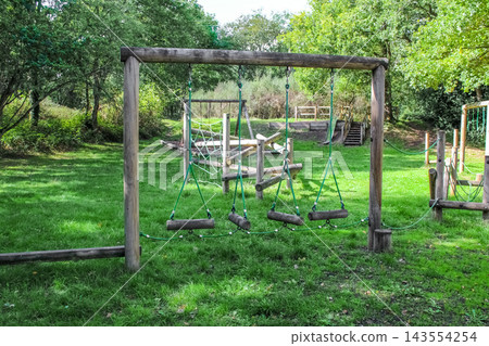 A woodland playground with rustic wooden climbing frames, ropes, and a nest swing sits on a lush green lawn, surrounded by dense trees and a picnic area under a leafy canopy in the soft daylight. A woodland playground with rustic wooden climbing frames, ropes, and a nest swing sits on a lush green lawn, surrounded by dense trees and a picnic area under a leafy canopy in the soft daylight. 143554254