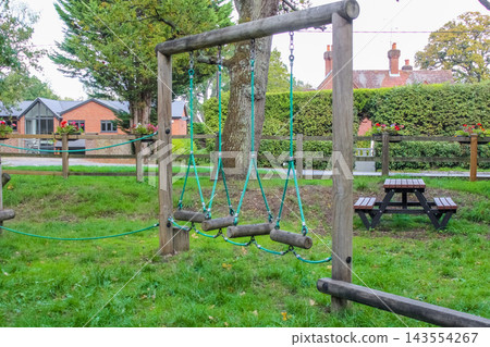 A rustic wooden playground nestled in a lush green forest. It features log climbing structures, rope nets, and swings, all enclosed by a timber fence under the dappled sunlight of leafy trees. A rustic wooden playground nestled in a lush green forest. It features log climbing structures, rope nets, and swings, all enclosed by a timber fence under the dappled sunlight of leafy trees. 143554267