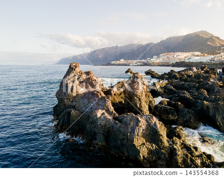 Jagged lava spires rise from the sea below distant cliffs. Sharp volcanic spires break the surface in the foreground. Behind them the town of Los Gigantes and its vast basalt cliffs recede into soft m Jagged lava spires rise from the sea below distant cliffs. Sharp volcanic spires break the surface in the foreground. Behind them the town of Los Gigantes and its vast basalt cliffs recede into soft m 143554368