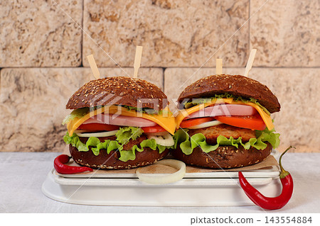 Two double cheeseburgers on whole-grain sesame seed buns featuring fresh lettuce, tomato, cream cheese, chili pepper, bacon, onion and a patty served on a wooden tray, food delivery concept, menu Two double cheeseburgers on whole-grain sesame seed buns featuring fresh lettuce, tomato, cream cheese, chili pepper, bacon, onion and a patty served on a wooden tray, food delivery concept, menu 143554884
