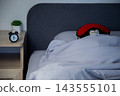 There is a black alarm clock on a wooden bedside table, and in the background a brown bear in a red mask sleeps wrapped in a white blanket under a gray headboard. Concept: the toy aesthetics of sleep 143555101