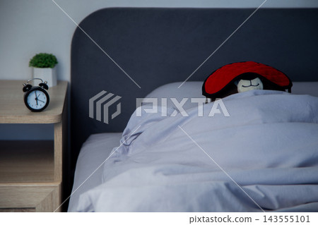 There is a black alarm clock on a wooden bedside table, and in the background a brown bear in a red mask sleeps wrapped in a white blanket under a gray headboard. Concept: the toy aesthetics of sleep There is a black alarm clock on a wooden bedside table, and in the background a brown bear in a red mask sleeps wrapped in a white blanket under a gray headboard. Concept: the toy aesthetics of sleep 143555101