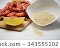 On a white background, there is a bowl of rice, a wooden board with frozen shrimp, a slice of lemon, and a few grains of cereal spilled onto the countertop. High quality photo 143555102