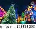 Illuminated Christmas tree flanked by buildings adorned with light projections in Alba, Italy. 143555205
